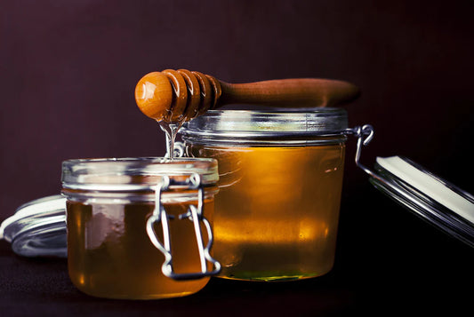 A jar of honey with a metal lid and handle, honey dripping from a wooden dipper into it.