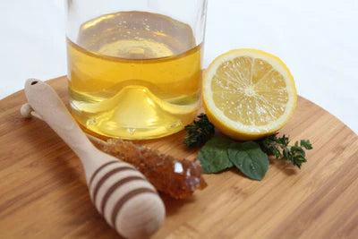 A wooden dipper, lemon halves, and honey on a table, with a glass of yellow liquid nearby.